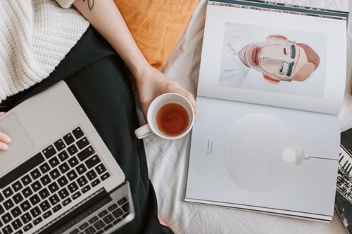 Free Top view of crop anonymous female in casual clothes with cup of tea using laptop while lying on bed with art book near Stock Photo