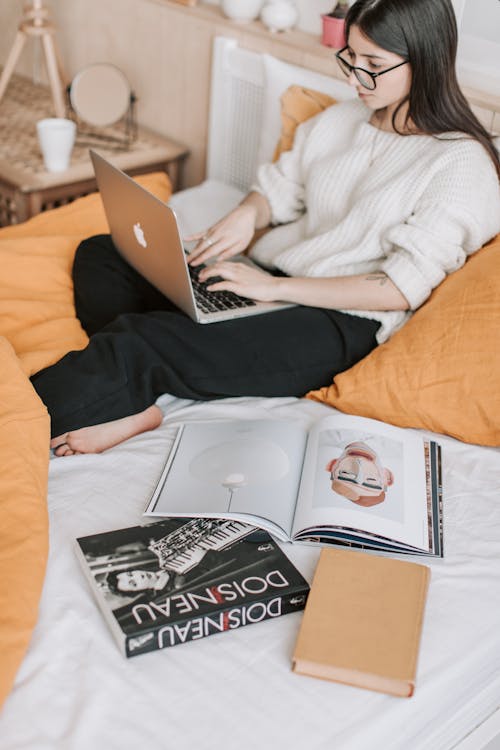 High angle of barefoot female in cozy sweater chilling on bed with blanket and books around while using laptop