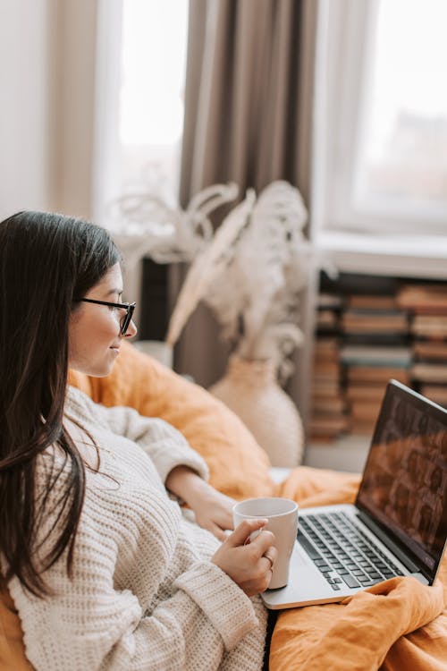 Side view of young content lady in warm white sweater and eyeglasses resting on cozy bed with mug of hot drink and browsing netbook in modern apartment in daytime