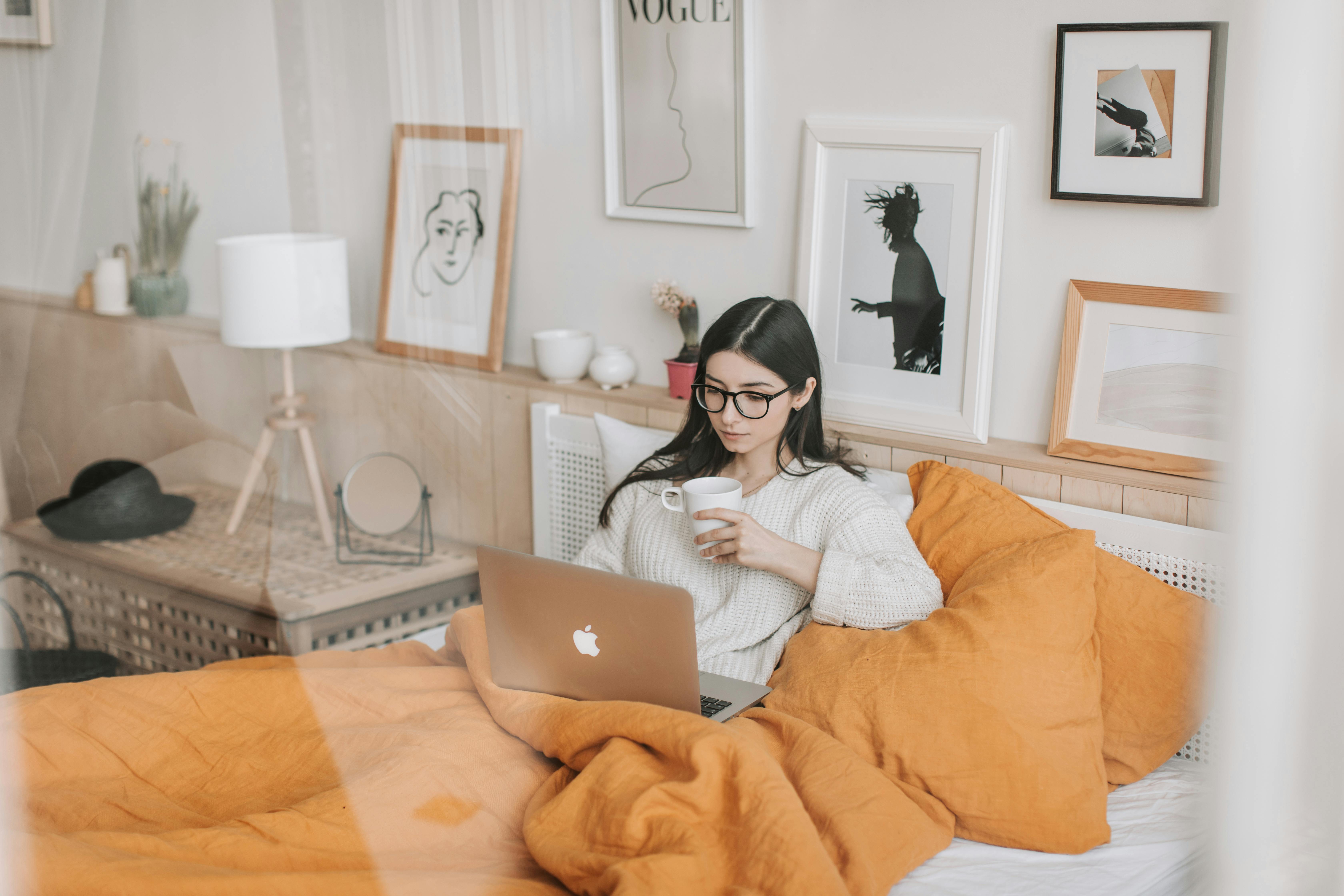 thoughtful woman with cup of drink browsing laptop in bed