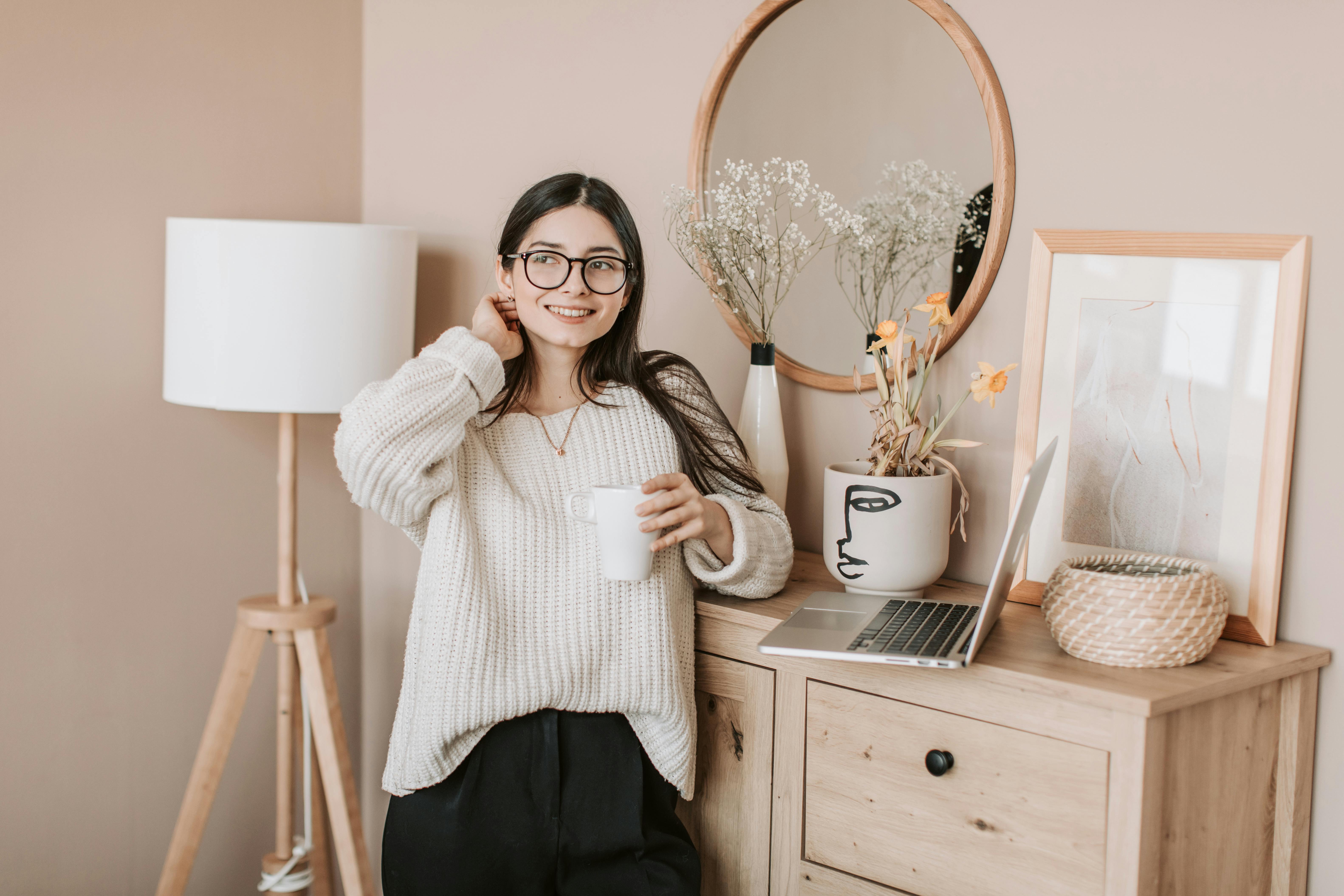 smiling woman with cup of drink and laptop in bedroom