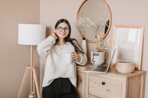 Dreamy smiling young lady in white sweater and eyeglasses with mug of hot beverage looking away happily while standing near bedroom dresser and browsing laptop
