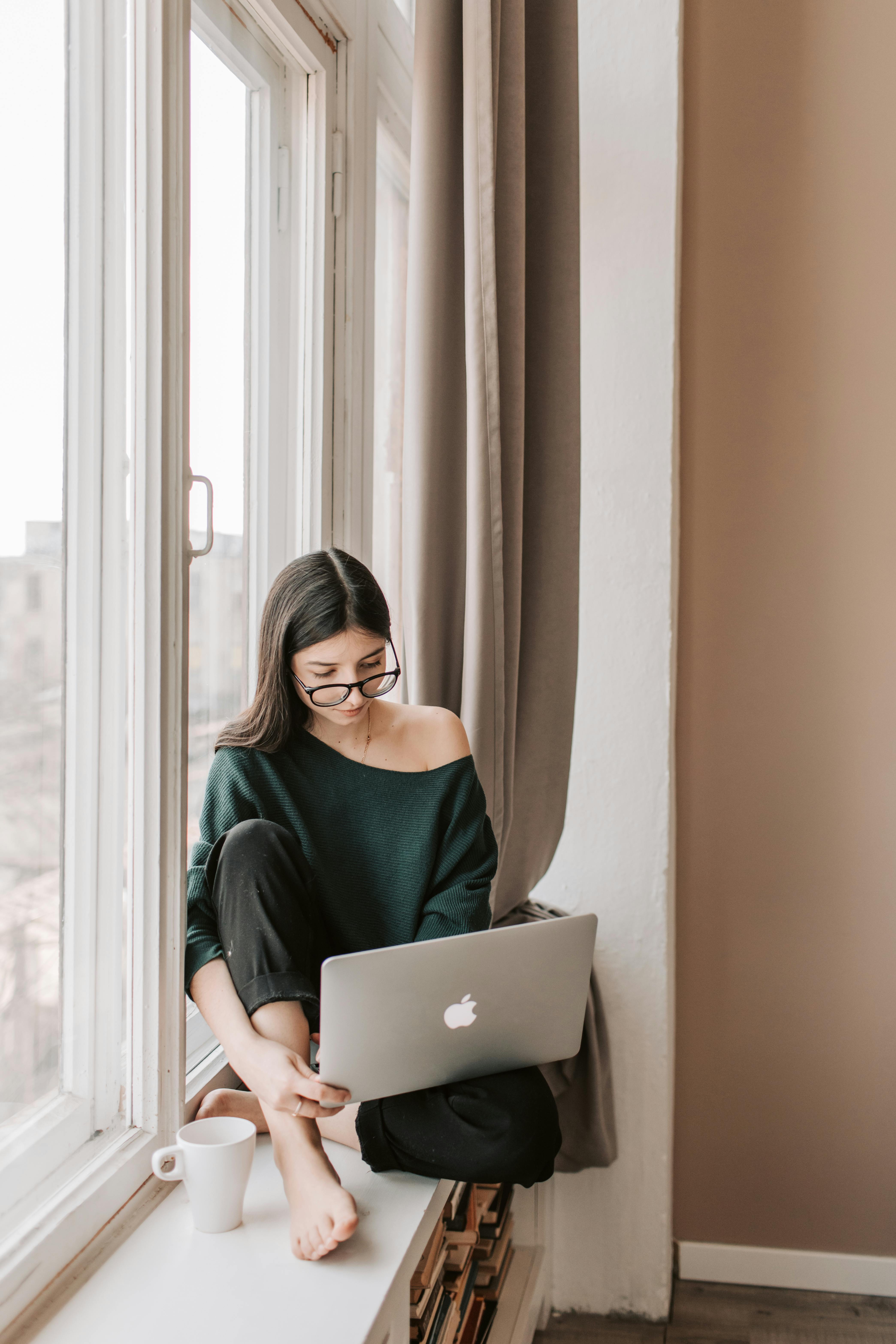young woman with mug using laptop on windowsill