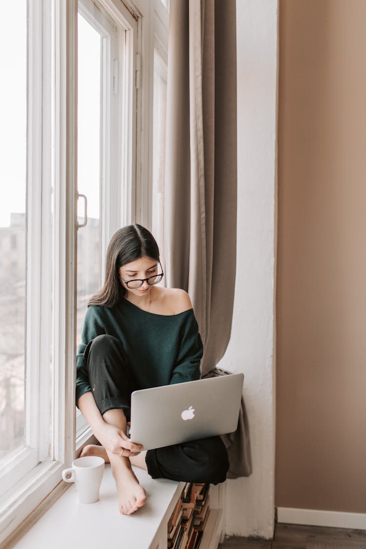 Young Woman With Mug Using Laptop On Windowsill