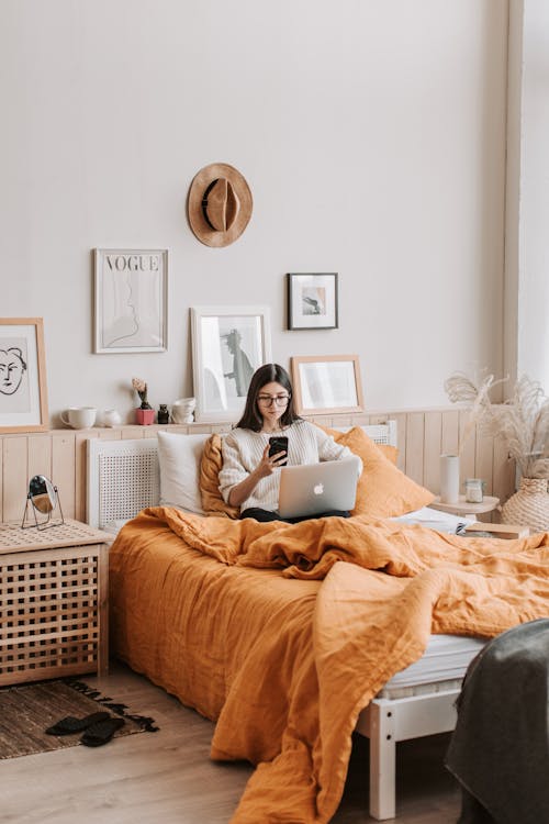 Full body of brunette in casual warm sweater text messaging via smartphone and surfing internet on netbook while resting on comfortable bed in morning