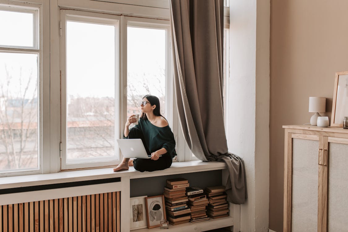 Free Young female in casual outfit sitting with cup of hot drink on windowsill and browsing netbook while looking out window pensively in cozy bedroom in late autumn Stock Photo