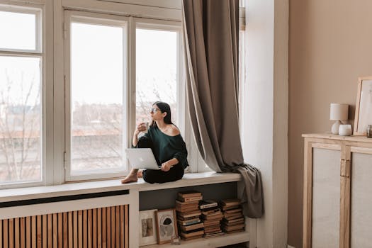 Young female in casual outfit sitting with cup of hot drink on windowsill and browsing netbook while looking out window pensively in cozy bedroom in late autumn