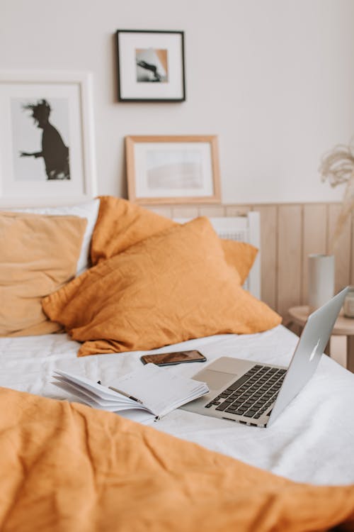 Comfortable disheveled empty bed with orange pillows and blanket with laptop and opened notebook on top near white wall decorated with paintings in daylight