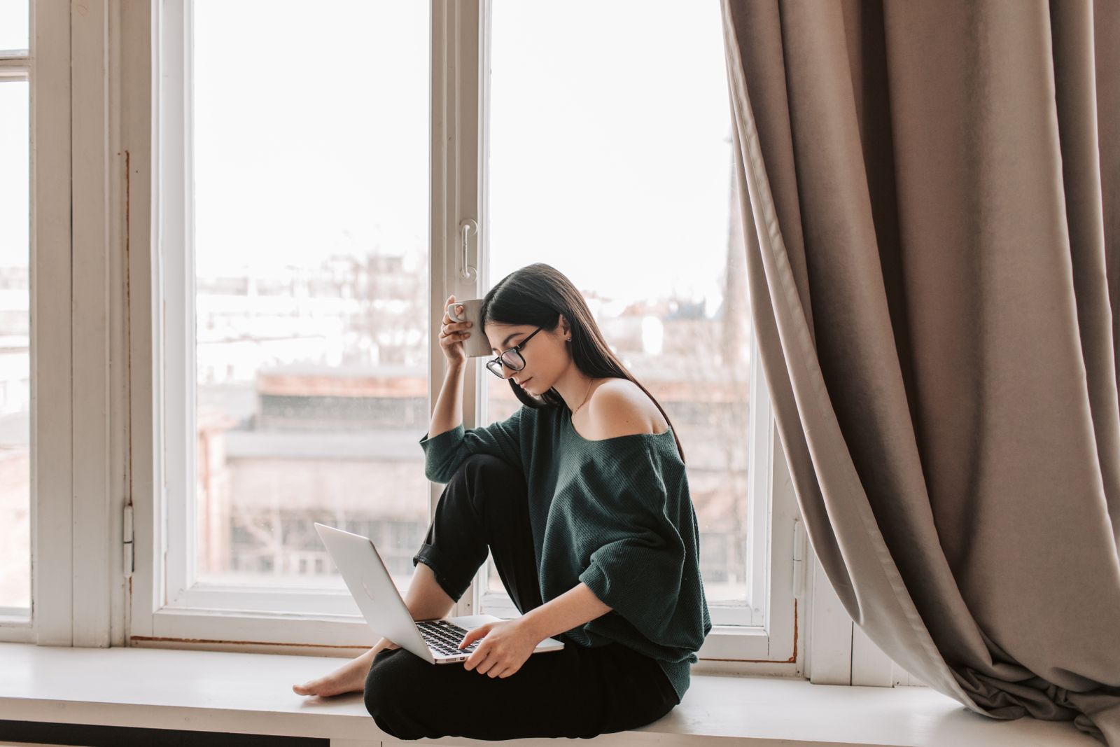 Woman wearing glasses sitting on a window sill working on a laptop with a warm drink