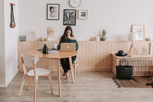 Free Full length of pensive brunette in eyeglasses sitting at table with legs crossed and working on laptop in cozy creative living room Stock Photo