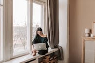 Young female freelancer sitting with laptop on windowsill at home