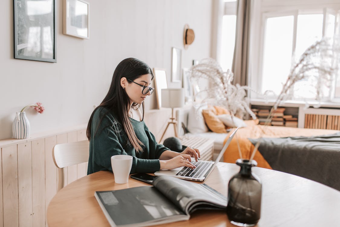 Free Side view of female remote worker in eyeglasses and casual wear typing on netbook while sitting at wooden table with cup and open magazine in bedroom at home Stock Photo