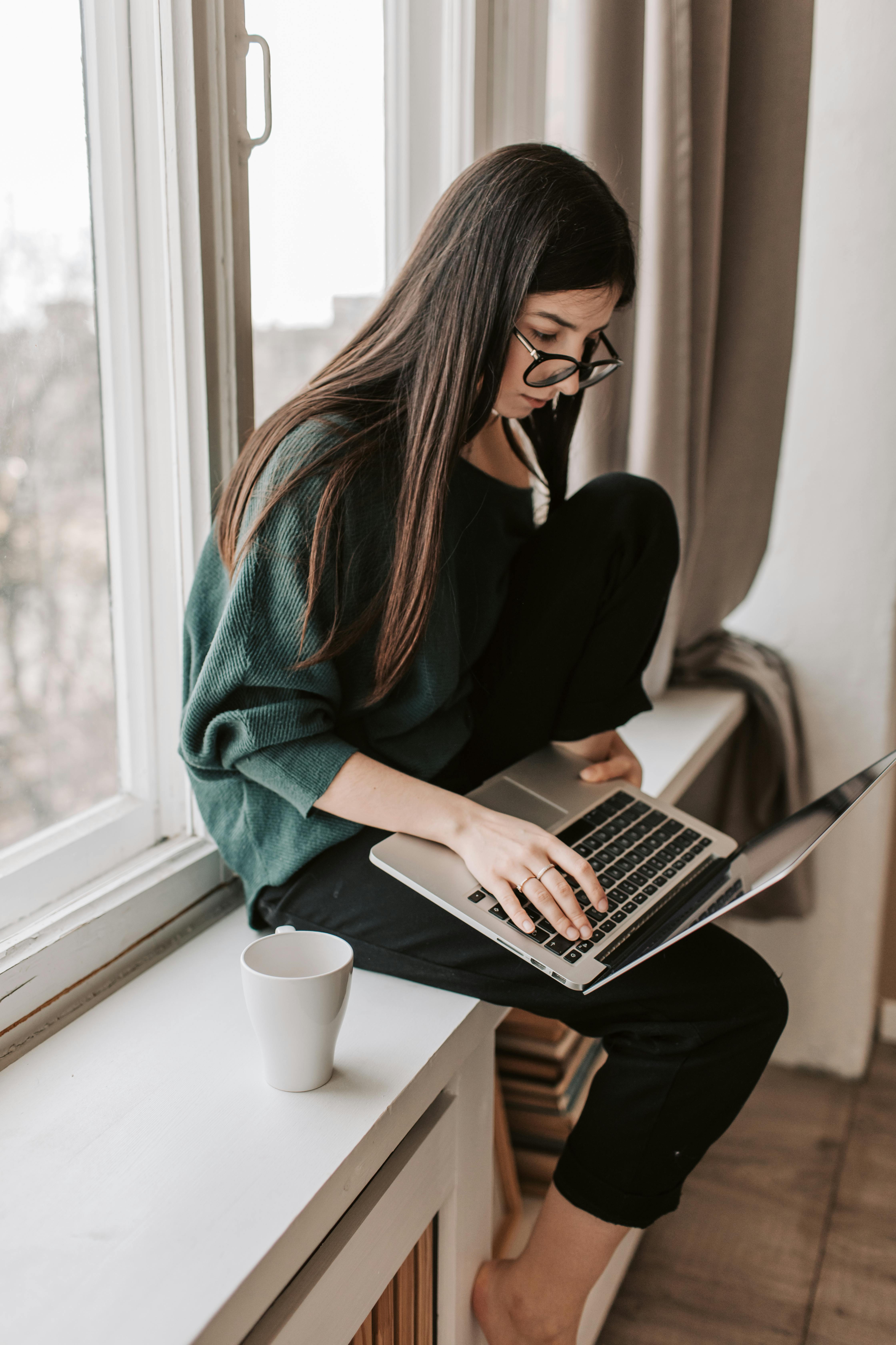 concentrated female freelancer typing on laptop on windowsill at home