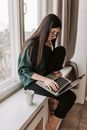 Side view of focused female remote worker in casual wear sitting on windowsill with netbook and cup of hot drink in flat in afternoon