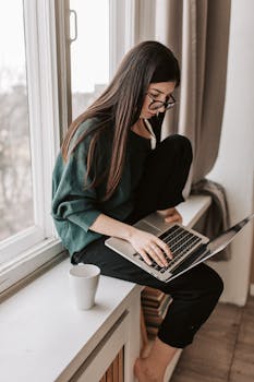 Side view of focused female remote worker in casual wear sitting on windowsill with netbook and cup of hot drink in flat in afternoon