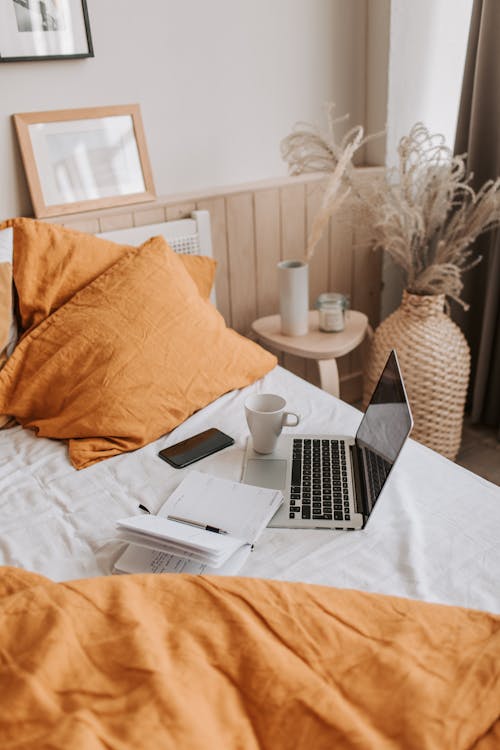 From above of open netbook with black screen and cup of coffee with smartphone and notebook placed on crumpled bed sheet in bedroom with big vase on floor
