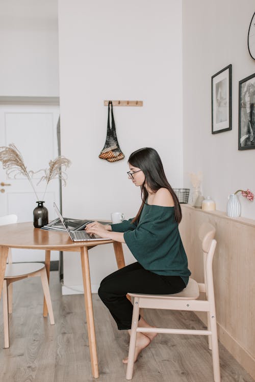 Side view of female freelancer in eyeglasses and casual wear working on netbook while sitting at wooden chair in light apartment