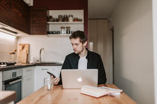 Young male remote employee in casual wear chatting on cellphone while sitting at wooden table in front of open netbook and copybook with glass of water in flat