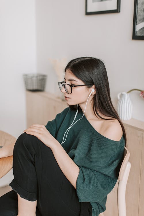 Side view of thoughtful young lady in eyeglasses and casual wear listening to music in earbuds while sitting on wooden chair near white wall with pictures and ceramic vase