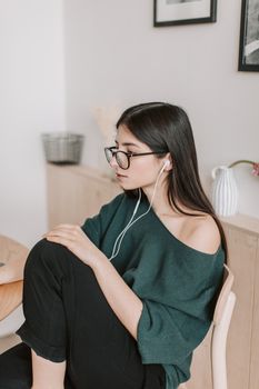 一位戴着眼镜、穿着休闲装的年轻女士，正坐在靠白墙的木椅上，旁边摆着图片和陶瓷花瓶，她侧身沉思
