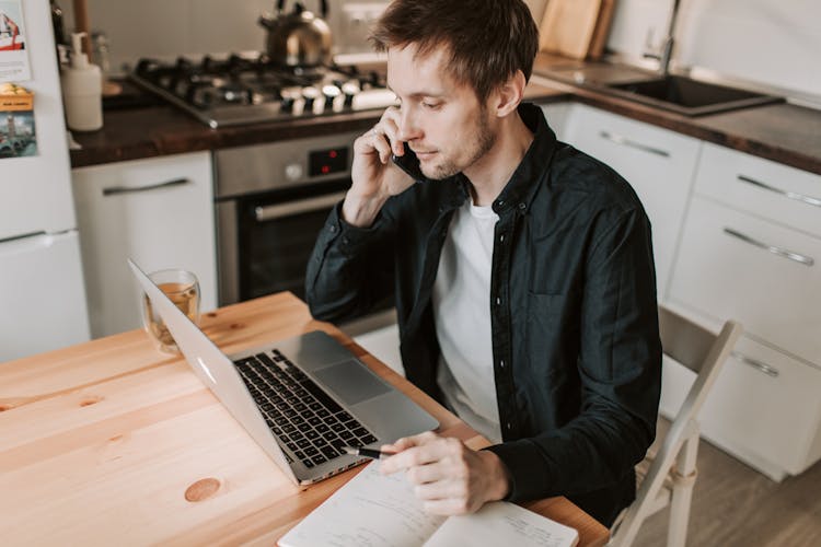 Male Freelancer Making Phone Call And Watching Laptop In Kitchen