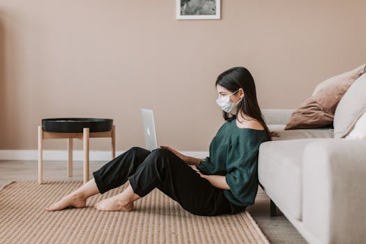 Side view of young female remote worker in casual wear and sterile mask sitting with netbook on rug leaning on sofa during COVID 19 pandemic