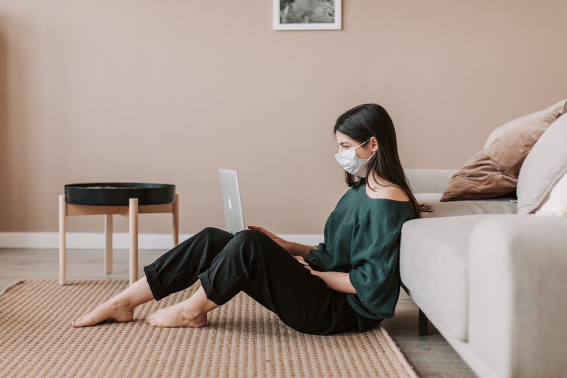 Free Side view of young female remote worker in casual wear and sterile mask sitting with netbook on rug leaning on sofa during COVID 19 pandemic Stock Photo