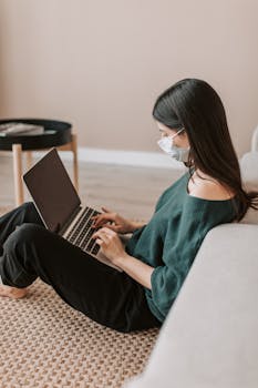 A woman wearing a mask works from home on a laptop in a comfortable living room setting.
