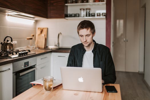 Pensive young male freelancer in casual clothes watching netbook while sitting at wooden table with glass of water and copybook in apartment