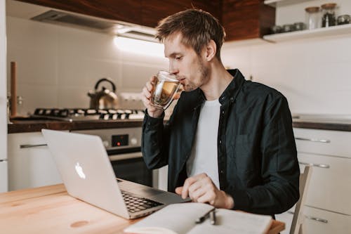 Free Young male freelancer in casual wear drinking tea while watching netbook and sitting at wooden table with open notebook in flat Stock Photo