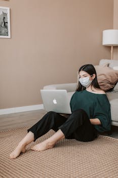 Young woman wearing mask working remotely on laptop in modern living room.