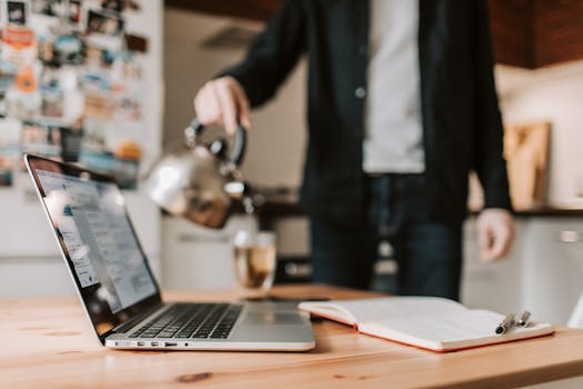A person preparing coffee while working remotely on a laptop in a cozy kitchen setup.