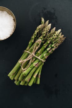 A fresh bundle of asparagus tied with twine on a dark surface, next to a bowl of sea salt.