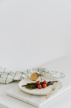 Minimalistic plate with asparagus, cherry tomatoes, and lemon on a white background.