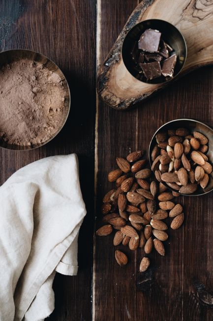 A wooden bowl of dark raw cacao powder next to a small pile of crunchy cacao nibs and a few whole cacao pods on a linen surface.