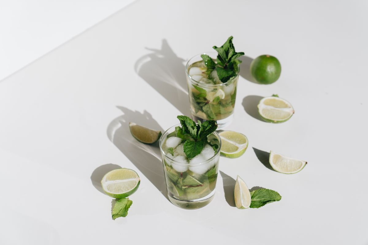 Two mojitos with fresh mint sprigs and lime pieces surrounded by lime wedges on white