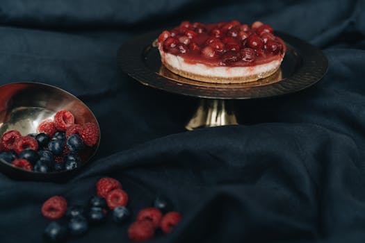 Close-up of a berry-topped cheesecake on a cake stand with raspberries and blueberries on a dark cloth.