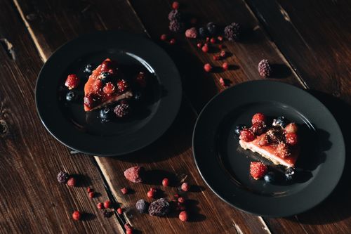 Free Two slices of berry cheesecake on black plates, styled with fresh berries on a rustic wooden table. Stock Photo