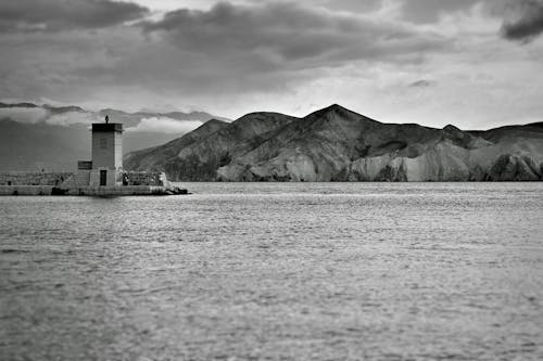 Black and white seascape with lonely lighthouse located against picturesque mountains on cloudy day