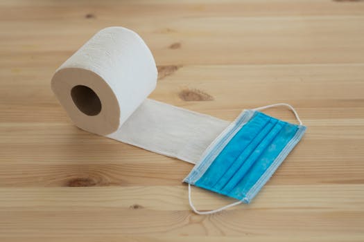A rolled toilet paper next to a face mask on a wooden table, symbolizing hygiene and safety during a pandemic.