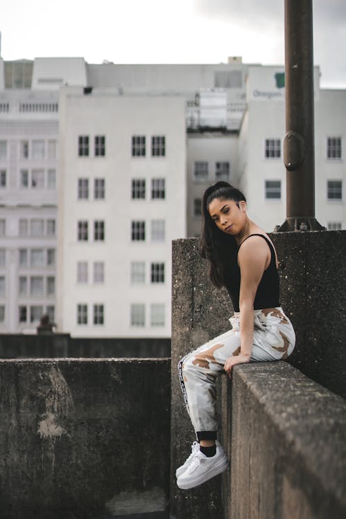 Woman in Black Tank Top and White Pants Sitting on Concrete Wall