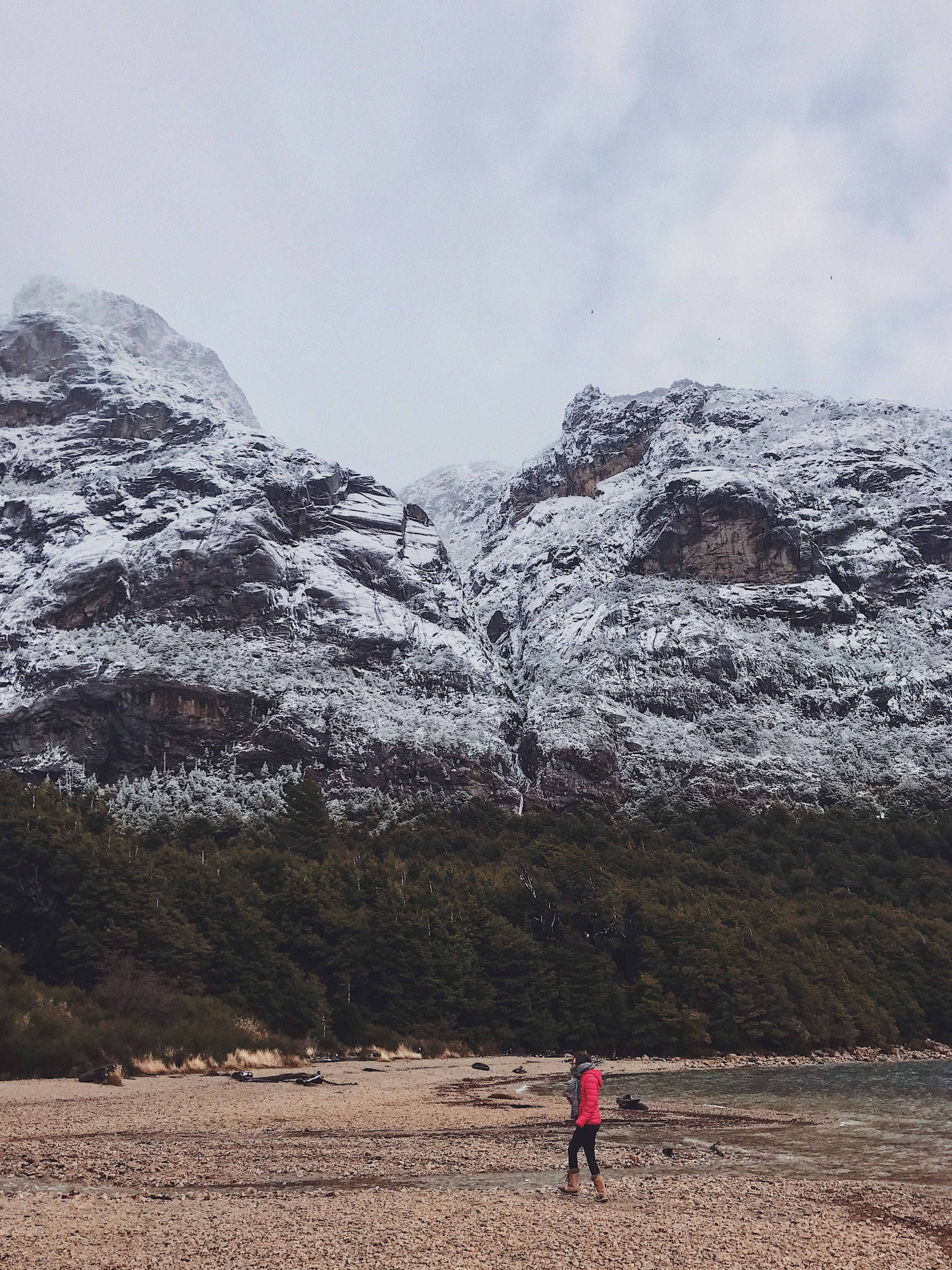 冬日白天，白雪皑皑的山脉和郁郁葱葱的森林间，一名徒步旅行者正行走在海滩上