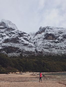 冬日白天，白雪皑皑的山脉和郁郁葱葱的森林间，一名徒步旅行者正行走在海滩上