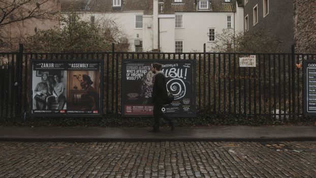 A man walks past posters on a cobblestone street lined with buildings.