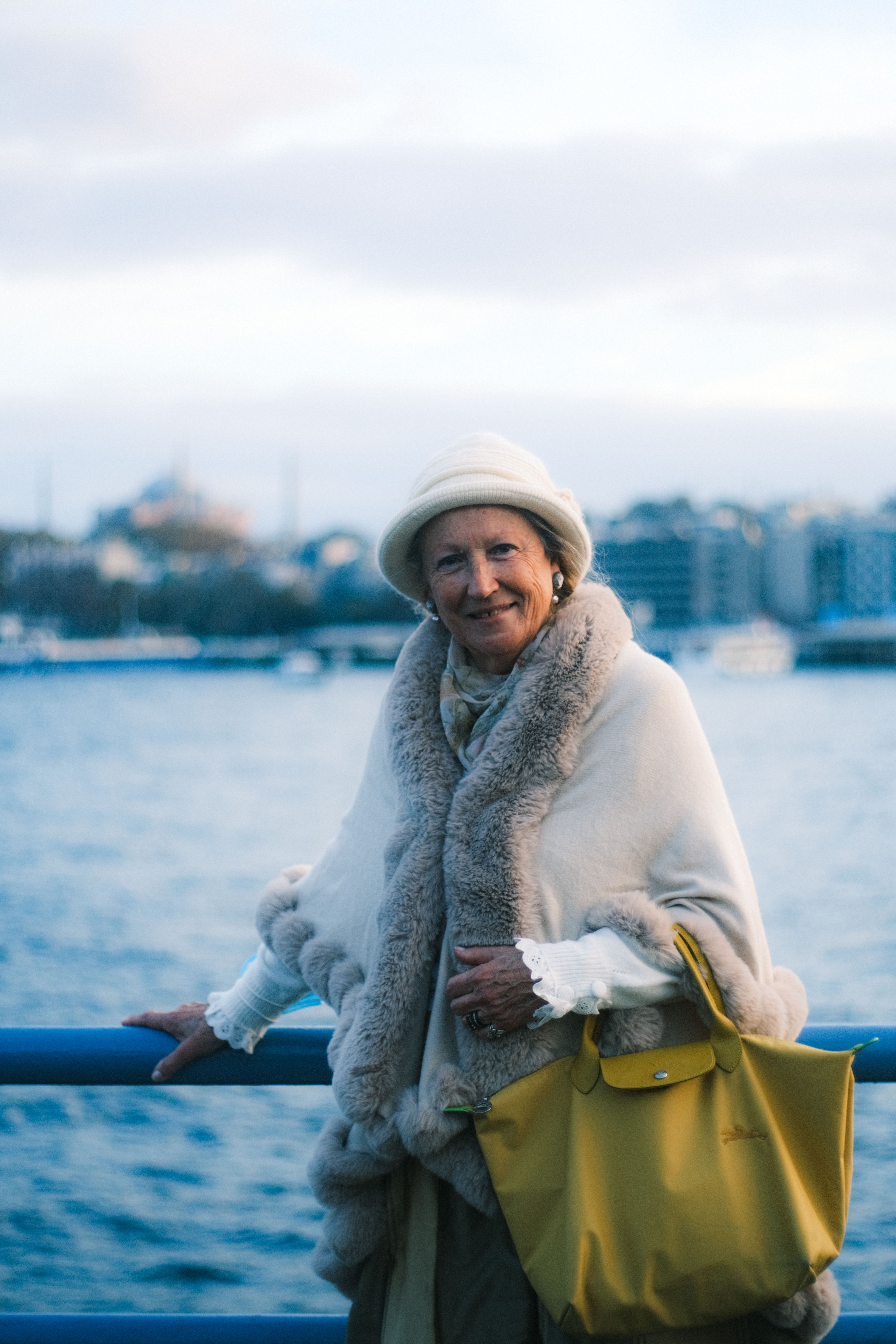 Senior woman standing by a waterfront in winter clothing, exuding elegance with cityscape in the background.