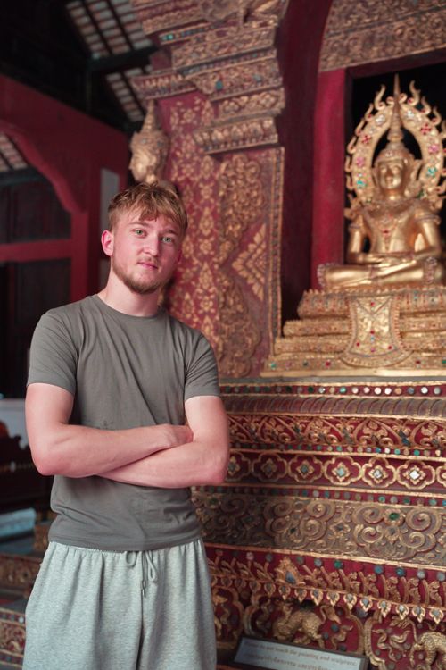 Free Young man standing beside an intricate golden Buddha statue in Chang Wat Chiang Mai, Thailand. Stock Photo