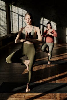 Two women practicing tree pose in a sunlit studio, focusing on wellness and balance.