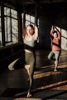 Two women practicing yoga in a serene sunlit room, focusing on balance and flexibility.