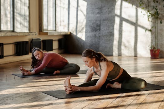 Two women practice yoga indoors, focusing on stretching and flexibility in a sunlit room.