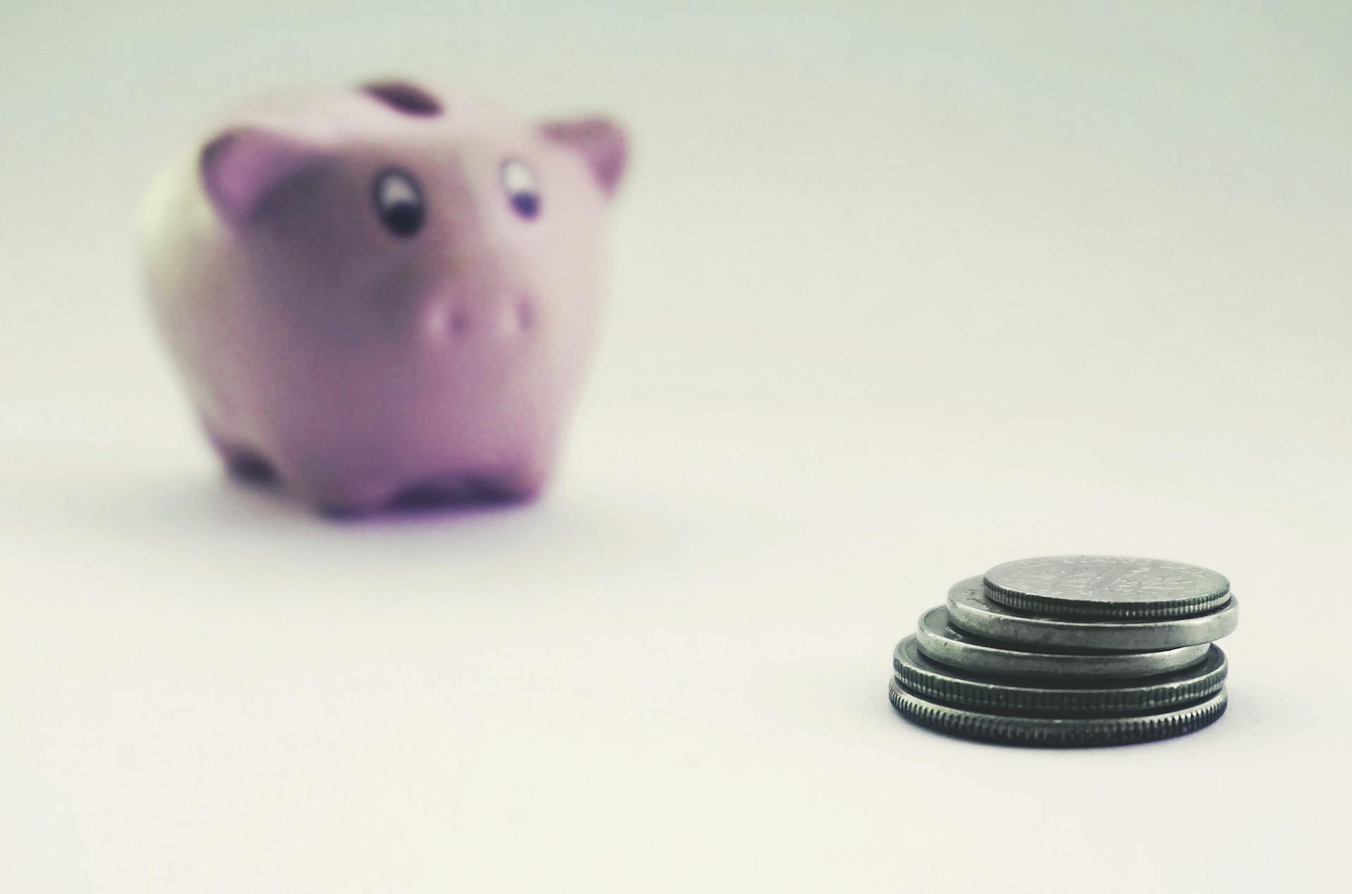 A pink piggy bank blurred in the background with stacked coins in the foreground on a white surface.